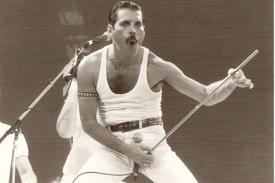 Sepia-toned image of Freddie Mercury mid-performance, wearing his iconic white tank top and pants with a studded belt, gripping a microphone stand and singing with intense energy. His expressive pose, open mouth, and commanding stance capture the electric atmosphere of a live Queen concert.
