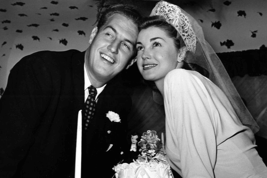Black and white photo of a joyful bride and groom leaning in close behind a wedding cake adorned with a floral topper and miniature figurines. The groom wears a dark suit with a patterned tie and boutonniere, while the bride smiles sweetly in a light gown and lace veil, both looking slightly off-camera.
