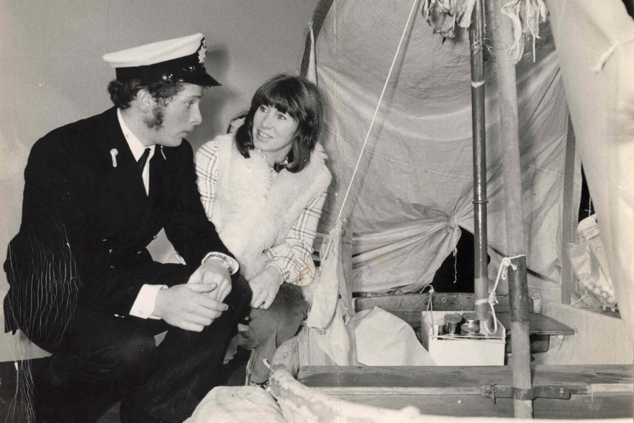 Black-and-white photo of a man in a naval uniform sitting and conversing with a woman in a plaid shirt and fur vest beside a small lifeboat display. The lifeboat is outfitted with makeshift sails, canned supplies, and survival gear, suggesting a museum or commemorative setting.