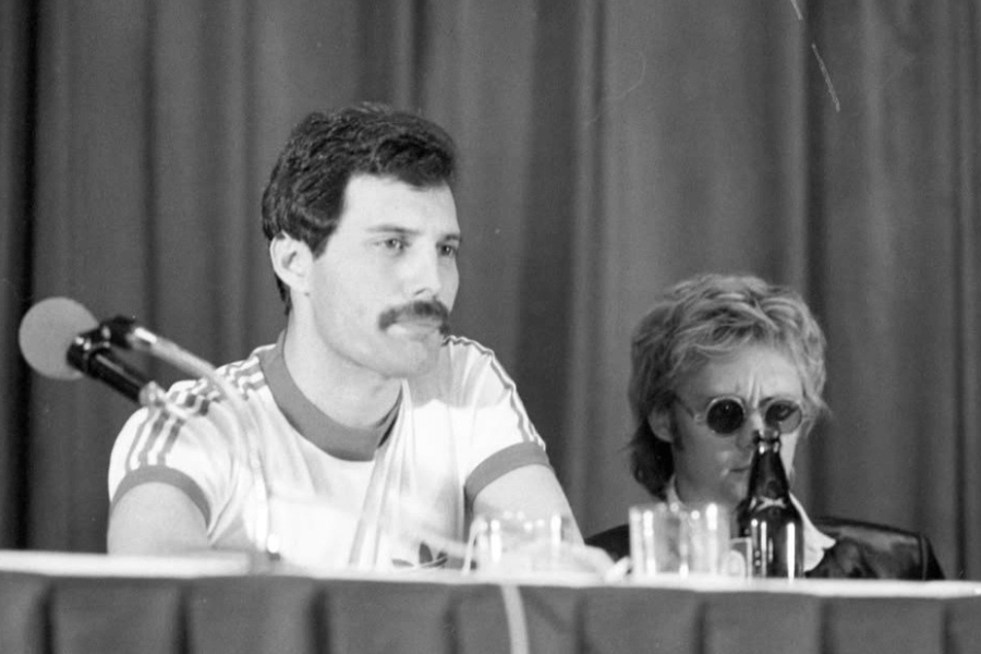 Black and white photo of Freddie Mercury seated at a press conference table, wearing a retro Adidas ringer t-shirt and looking serious, with a microphone in front of him. Beside him is Queen drummer Roger Taylor in sunglasses, partially obscured by a beer bottle, both sitting against a curtain backdrop.