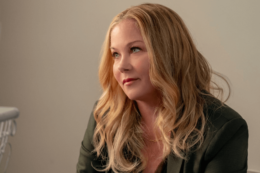 A woman with long, wavy blonde hair and subtle makeup sits indoors in a softly lit room, wearing a dark green blazer. She gazes to the side with a gentle, thoughtful expression.