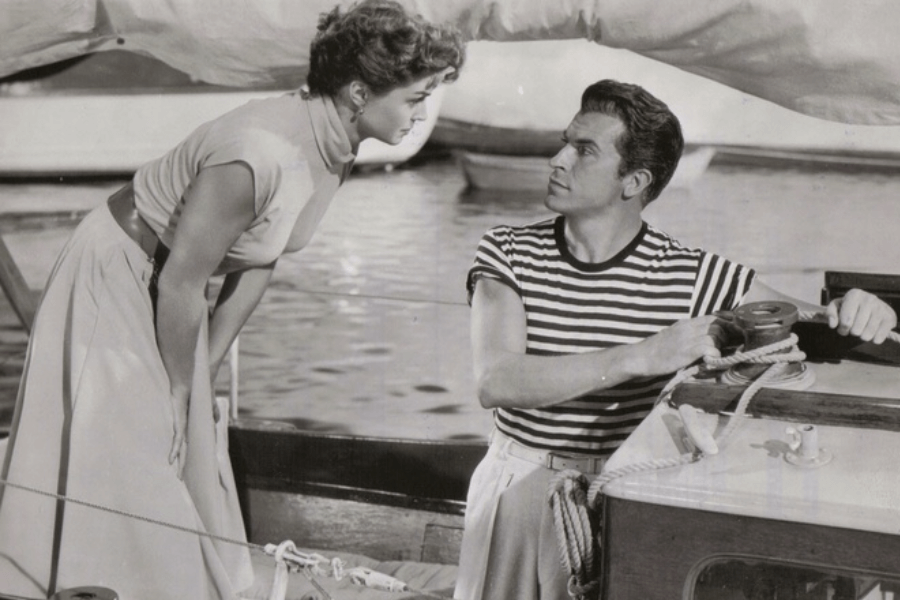 Black and white photo of a woman in a sleeveless blouse and high-waisted skirt leaning toward a man aboard a sailboat. The man, wearing a striped shirt and belted trousers, sits at the helm, gripping the rigging as they exchange a serious gaze. Calm water and other boats are visible in the background.