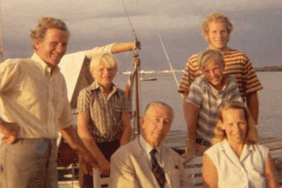 Faded color photo of a smiling group of seven people, including three adults and four children or teens, gathered on a dock beside a sailboat. The sky is overcast but warm-toned, and the group appears relaxed and happy, dressed in casual and semi-formal attire.