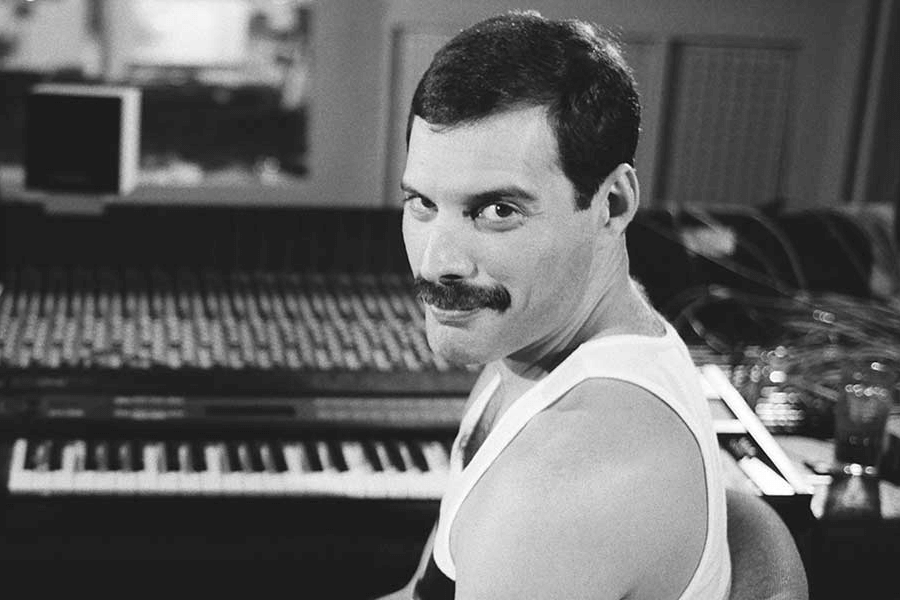 Black and white photo of Freddie Mercury sitting at a mixing console in a recording studio, turning to look over his shoulder with a confident smile. He wears a sleeveless white tank top, and the background features a keyboard and rows of audio equipment, capturing an intimate moment in the creative process.