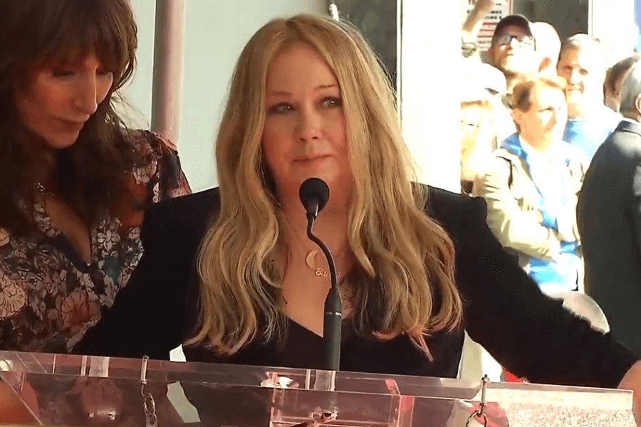 A woman with long blonde hair stands behind a podium with a microphone, delivering an emotional speech. Dressed in black, she is supported by another woman in a floral dress beside her, while a crowd watches in the background.