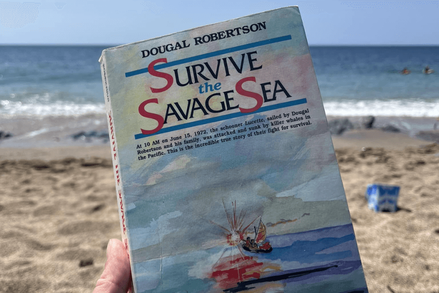 Close-up of a person holding a copy of Survive the Savage Sea by Dougal Robertson on a sandy beach with ocean waves in the background. The book cover features watercolor artwork of a lifeboat at sea firing a flare, and a blurb describing the true story of the Robertson family’s survival after their schooner was sunk by killer whales.