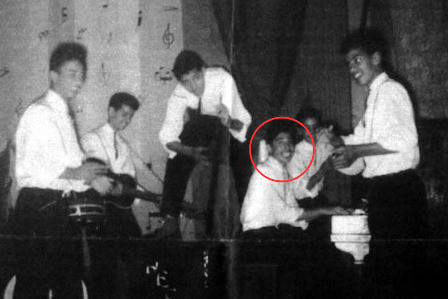 Grainy black and white photo of a teenage band performing on stage, with five young men dressed in white shirts and dark pants. One member, circled in red, is seated at a piano, smiling at the camera while the others play instruments or gather around playfully.