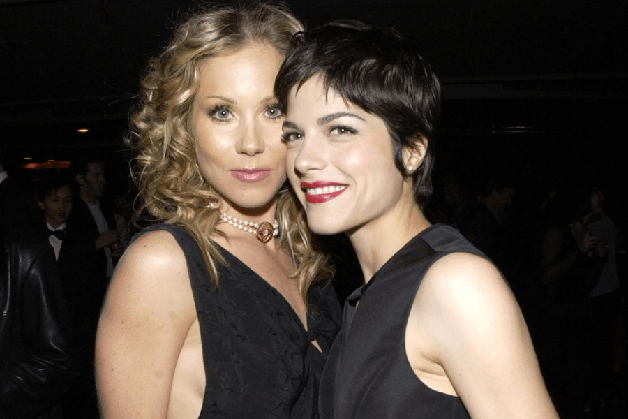 Two women pose closely at a formal event, both dressed in elegant black attire. One has curly blonde hair and wears a pearl necklace, while the other has a pixie haircut and red lipstick, smiling brightly as they enjoy the evening.