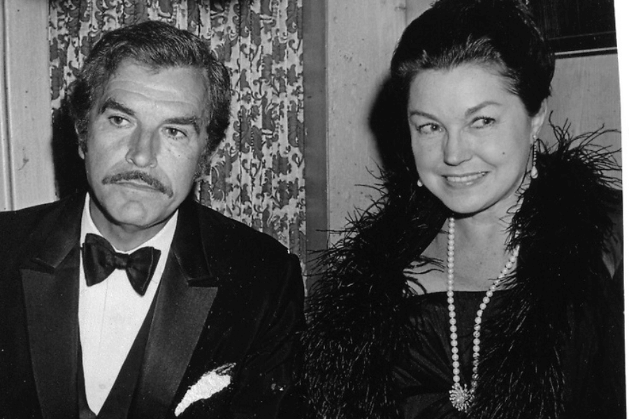 Black and white photo of a formally dressed couple at an indoor event. The man wears a classic tuxedo and bow tie with a serious expression, while the woman smiles in a strapless gown adorned with a long pearl necklace, feathered stole, and drop earrings. They appear to be seated together at a function.