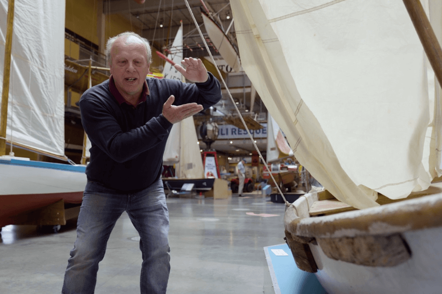 A man with light hair, wearing a dark sweater and jeans, enthusiastically gestures while explaining something beside a small, aged lifeboat on display in a maritime museum. Behind him are other sailboats and maritime exhibits, including a large "RESCUE" sign in the background.