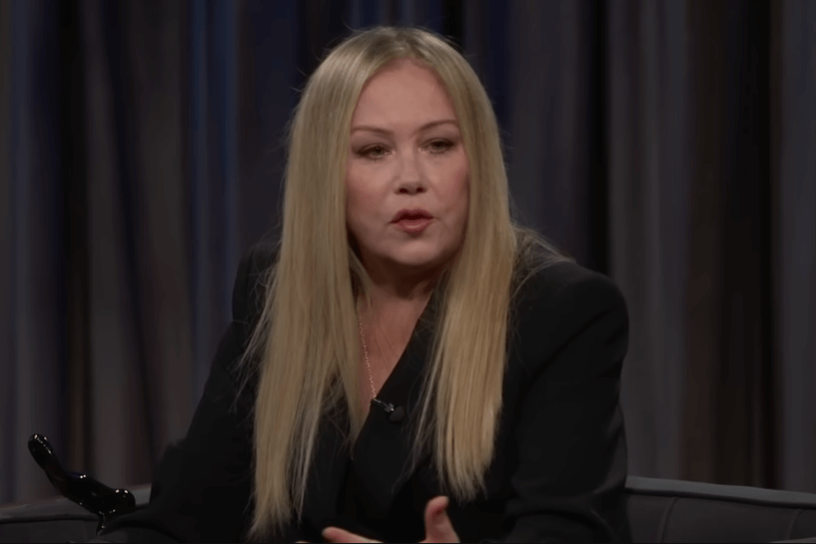 A woman with long, straight blonde hair and wearing a black blazer speaks while seated on a talk show set. She gestures with her hands during the conversation, with dark curtains forming the backdrop.
