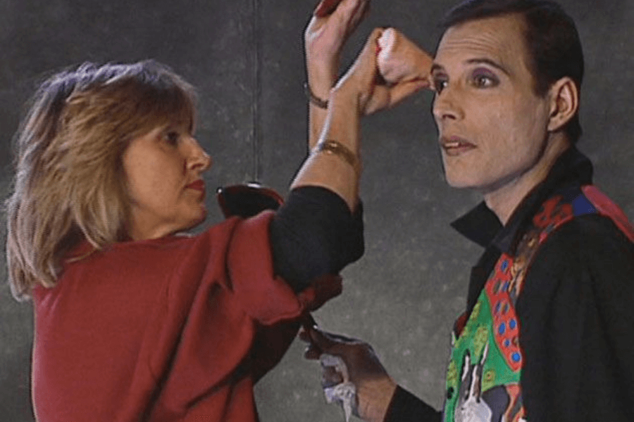 He wears his iconic cat-themed vest and looks off-camera with a calm expression as the makeup artist applies product near his eye, capturing a rare and intimate moment from his final public appearance.