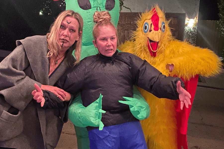 Three people pose in humorous Halloween costumes at night: one with dramatic face paint and a cloak, another in an inflatable alien abduction suit with a bewildered expression, and the third in a bright yellow chicken costume with exaggerated features.