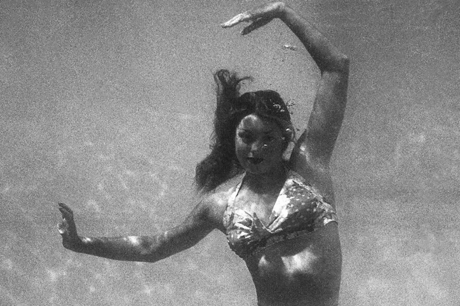 Grainy black and white underwater photo of a woman in a patterned bikini, striking a graceful pose with one arm curved above her head and the other extended outward. Her hair floats freely around her, and bubbles trail near her face as she performs an elegant move beneath the surface.