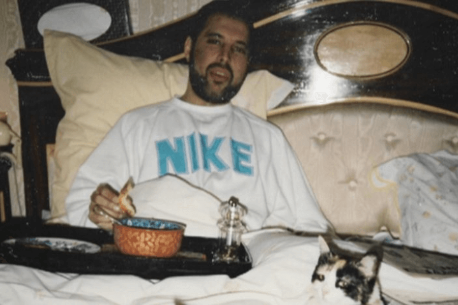 A frail Freddie Mercury sits up in bed wearing a white Nike sweatshirt, eating from a breakfast tray while a calico cat rests at his side. The photo captures a quiet and intimate moment during his final months, with soft lighting and a peaceful domestic setting.