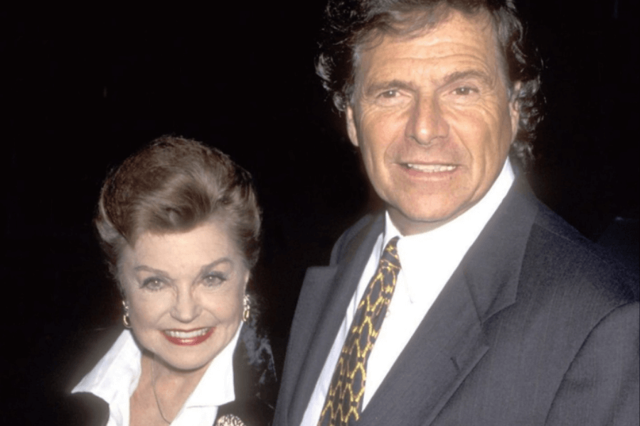 Color photo of an elegantly dressed older woman and man posing together at an event. The woman smiles warmly, wearing a white-collared black jacket with gold earrings and a brooch, while the man, also smiling, wears a dark suit with a white shirt and patterned tie. They appear to be attending a formal gathering.