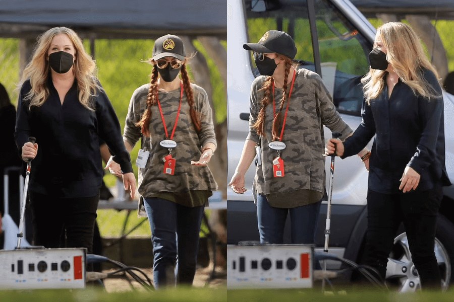 Side-by-side images show a woman with long blonde hair wearing a black mask and dark outfit walking with the aid of a cane, accompanied by a crew member in a camo shirt, braids, and a baseball cap. They appear to be on a production set with vans and equipment in the background.