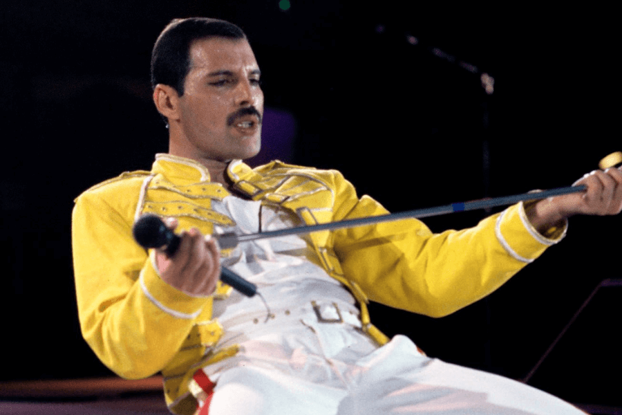 Freddie Mercury performing live on stage, leaning back while gripping a microphone stand with intensity. He wears his iconic yellow military-style jacket and white pants with red and gold accents, captured mid-song in one of Queen's most legendary concert moments.