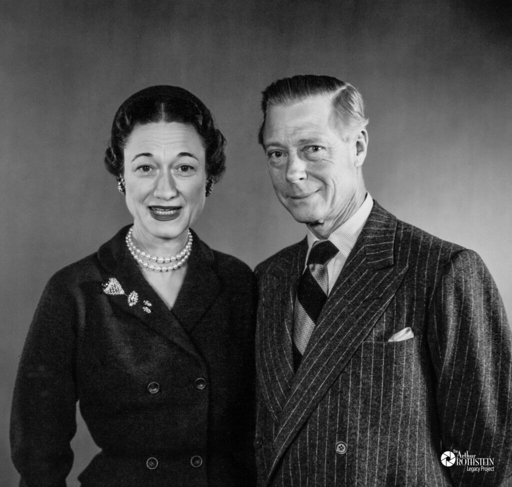 A black-and-white portrait of a smiling couple in formal attire. The woman wears a pearl necklace, earrings, and a brooch on her dark coat; the man wears a pinstripe suit with a tie and pocket square.