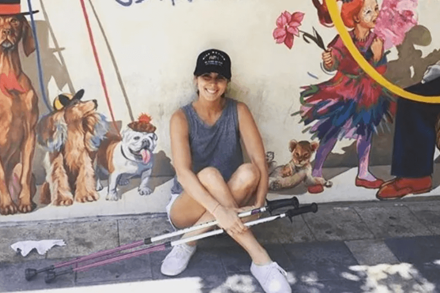 A smiling woman in a sleeveless top, shorts, and a black cap sits cross-legged on a sidewalk in front of a colorful mural featuring dogs in hats and circus-themed characters. She holds two walking poles across her legs.