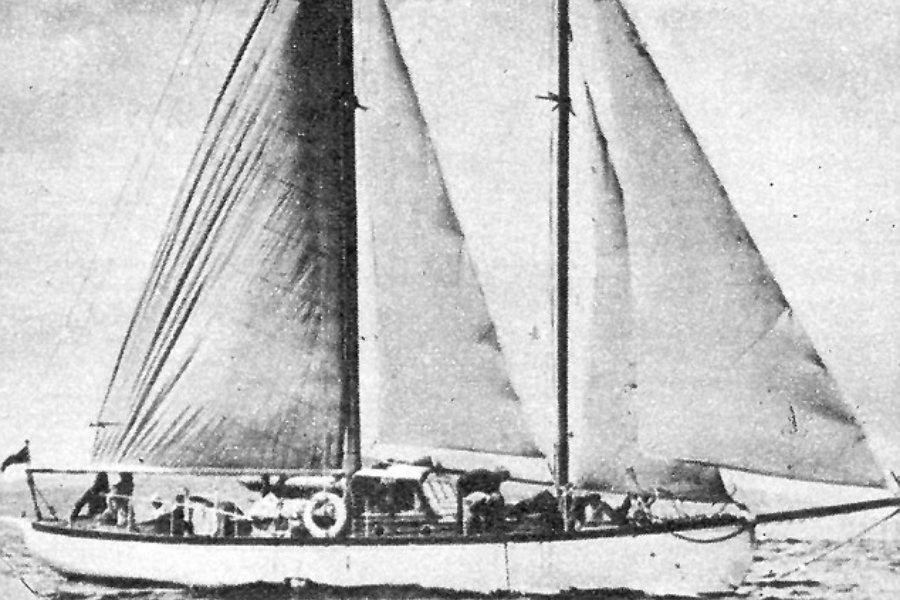 Grainy black-and-white image of a mid-sized sailboat with two raised sails, gliding across open water. Several people are visible onboard, surrounded by rigging and deck gear, with a lifebuoy mounted on the side railing and a flag flying at the stern.