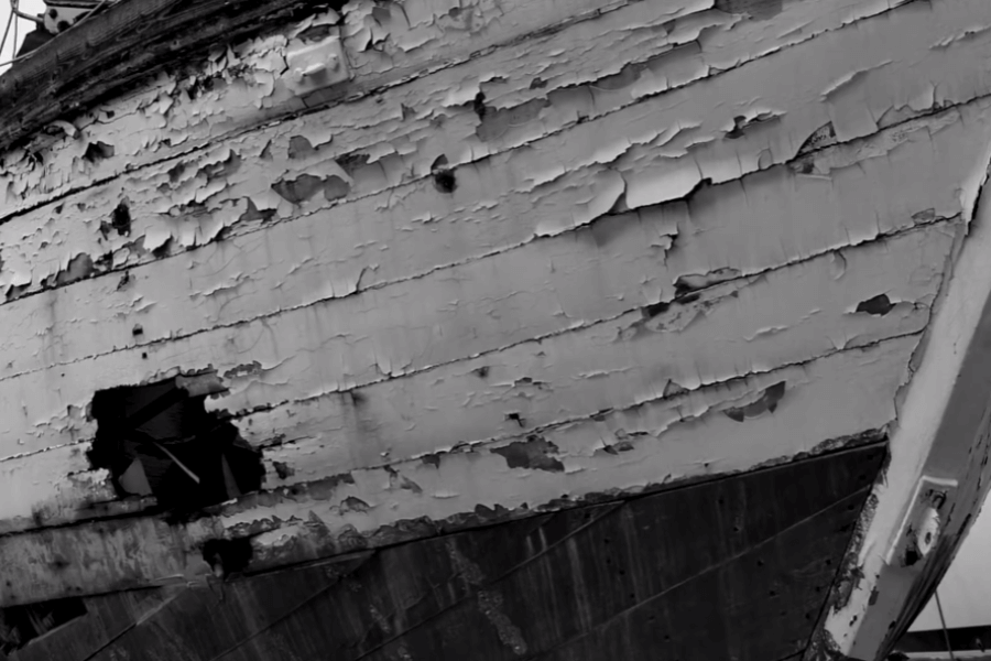 Close-up black-and-white image of a heavily weathered wooden boat hull with peeling paint, deep cracks, and a large hole exposing interior damage. The surface shows extensive signs of neglect and decay, suggesting long-term exposure to harsh elements.