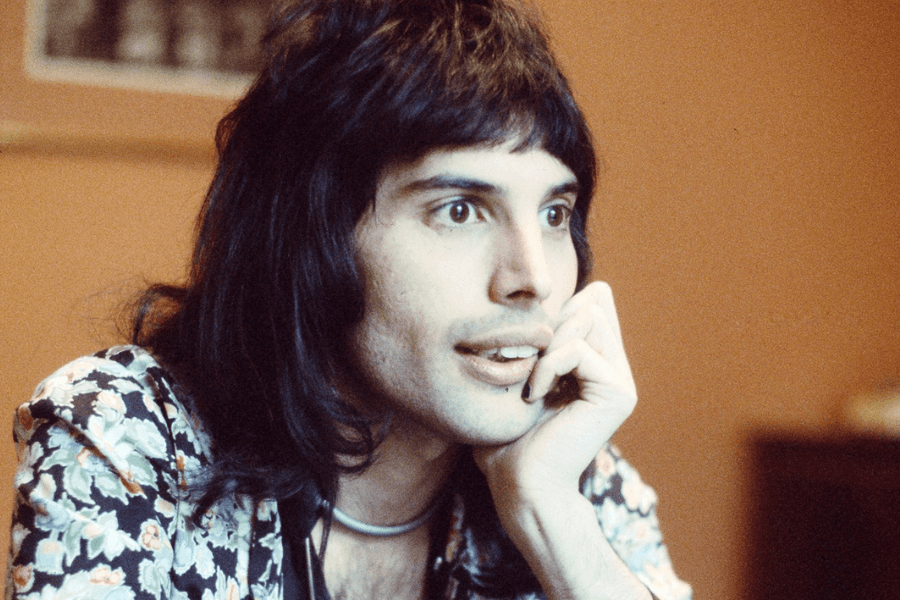 Candid photo of a young Freddie Mercury resting his face on his hand, gazing wide-eyed and thoughtfully into the distance. He has long dark hair, wears a floral shirt, and has black nail polish on one finger, capturing a soft and introspective moment.