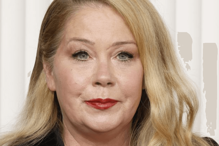 Close-up of a woman with long, wavy blonde hair and bold red lipstick, wearing subtle eye makeup and a black outfit, standing in front of a white background with vertical lines.