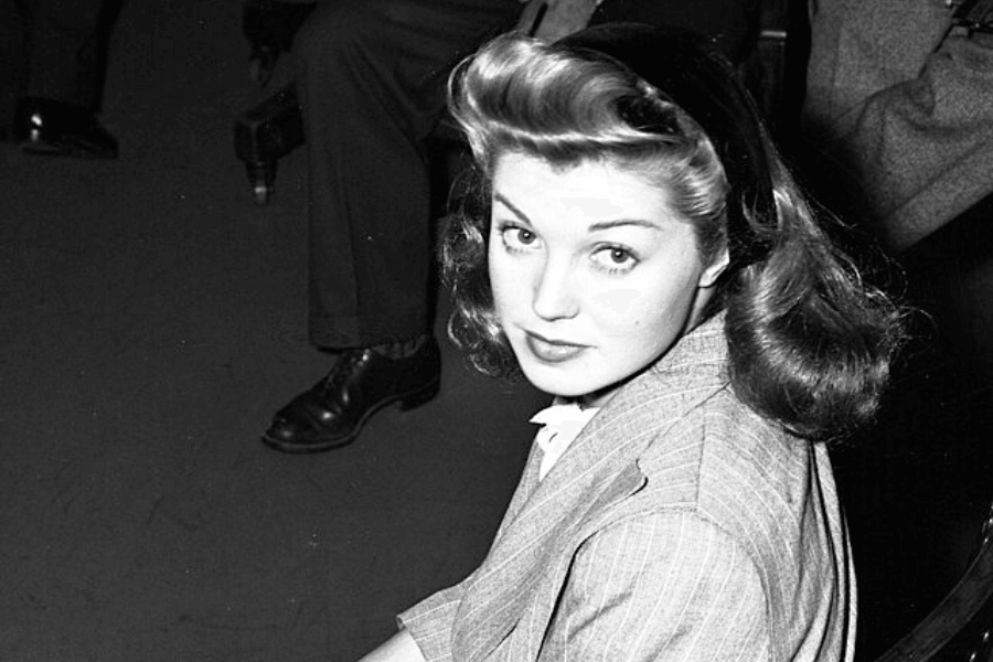 Black and white candid photo of a young woman with voluminous, styled hair and a dark headband, glancing up toward the camera. She’s seated, wearing a pinstripe jacket, surrounded by people in suits, likely in a public or formal setting.