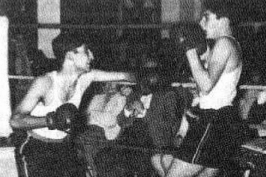 Grainy black and white photo of two young men in a boxing ring mid-match, both wearing tank tops, boxing gloves, and shorts. One fighter is in a defensive stance, while the other throws a punch, with spectators blurred in the background.