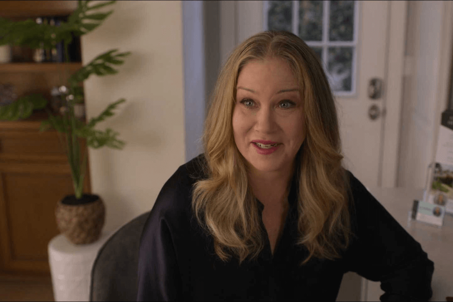 A woman with long, wavy blonde hair and a black blouse smiles while sitting indoors in a well-lit room. Behind her is a potted plant, a white door with glass panes, and a countertop with brochures.