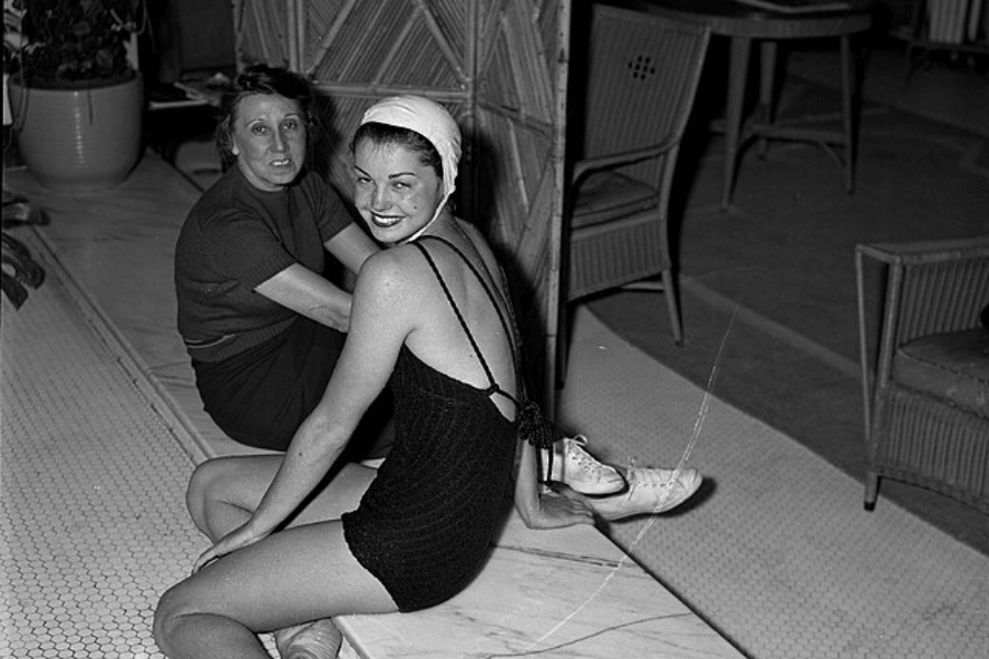 Black and white photo of a smiling woman in a textured one-piece swimsuit, swim cap, and white lace-up shoes, sitting poolside with another woman. The setting includes wicker furniture and tile flooring, suggesting an indoor pool or leisure area.