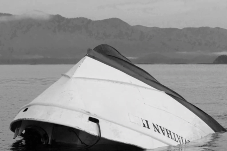 Black-and-white photo of an overturned boat partially submerged in calm water, with the name “LIVATHAN II” visible on the hull. Misty mountains loom in the background beneath a cloudy sky.