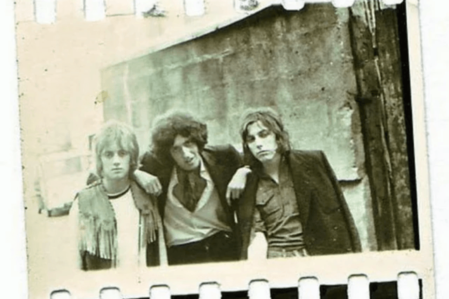 Faded, vintage-style photograph of three young men posing in an alleyway with worn brick walls. The man in the middle leans his arms over the shoulders of the two others, creating a casual, band-like pose. All three have long hair and 1970s rock-inspired clothing, giving off a gritty, early band photo vibe.