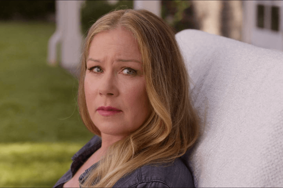A woman with long, blonde hair sits on a white chair outdoors, looking slightly to the side with a serious expression. The background includes green grass and part of a white building structure.
