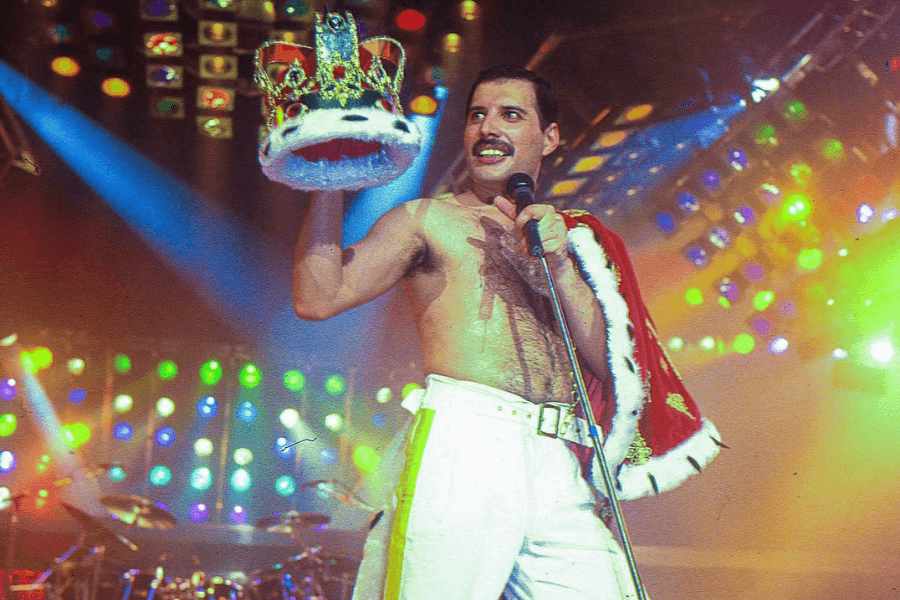 Freddie Mercury performing on stage in a dramatic moment, shirtless and draped in a red and white royal cape, holding a crown in one hand and a microphone in the other. He stands under vibrant, multicolored stage lighting with a backdrop of drums and spotlights, embodying his iconic regal stage persona.