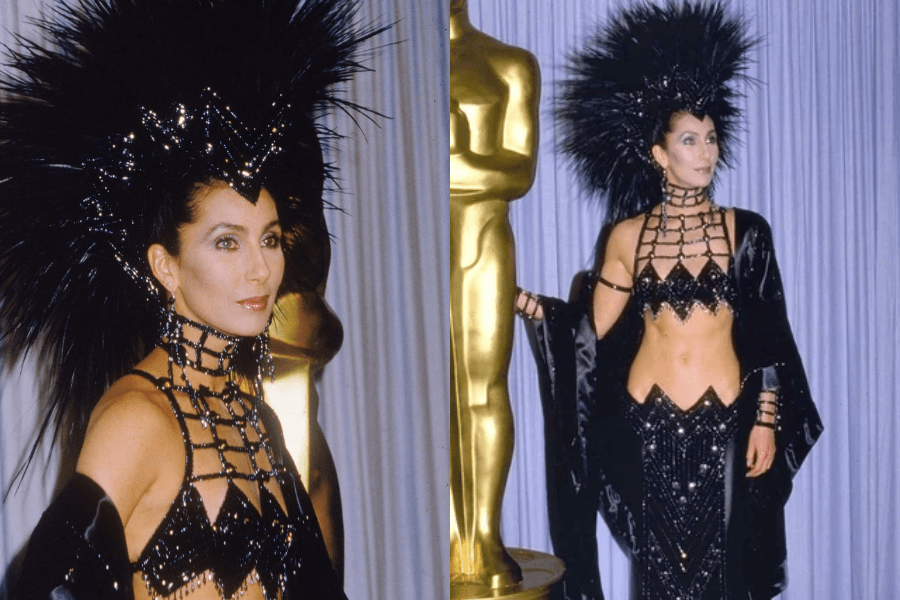 Cher poses at an awards show in a dramatic black outfit featuring a beaded bralette, matching high-waisted skirt with a zigzag hemline, and a long black shawl. She wears an elaborate feathered and bejeweled headpiece resembling a crown, standing next to a gold Oscar statue against a curtain backdrop.