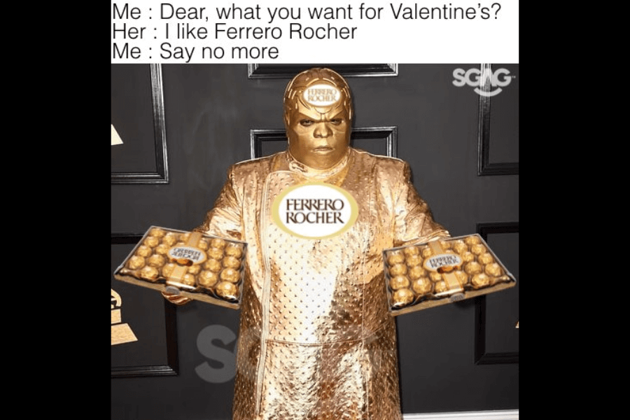 A meme shows a man fully dressed in metallic gold attire at an awards event, edited to resemble a Ferrero Rocher chocolate. He holds two large boxes of the chocolates, with logos also edited onto his chest and forehead. Above the image, a caption reads: “Me: Dear, what you want for Valentine’s? Her: I like Ferrero Rocher. Me: Say no more.”