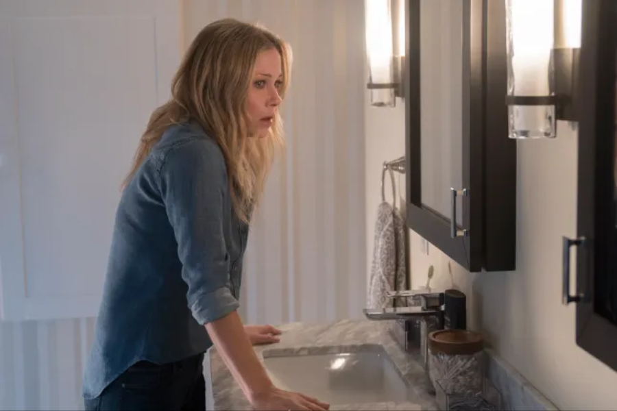 A woman with long blonde hair and a denim shirt leans over a bathroom sink, gazing into the mirror with a tense, emotional expression. The softly lit scene reflects a moment of introspection or distress.