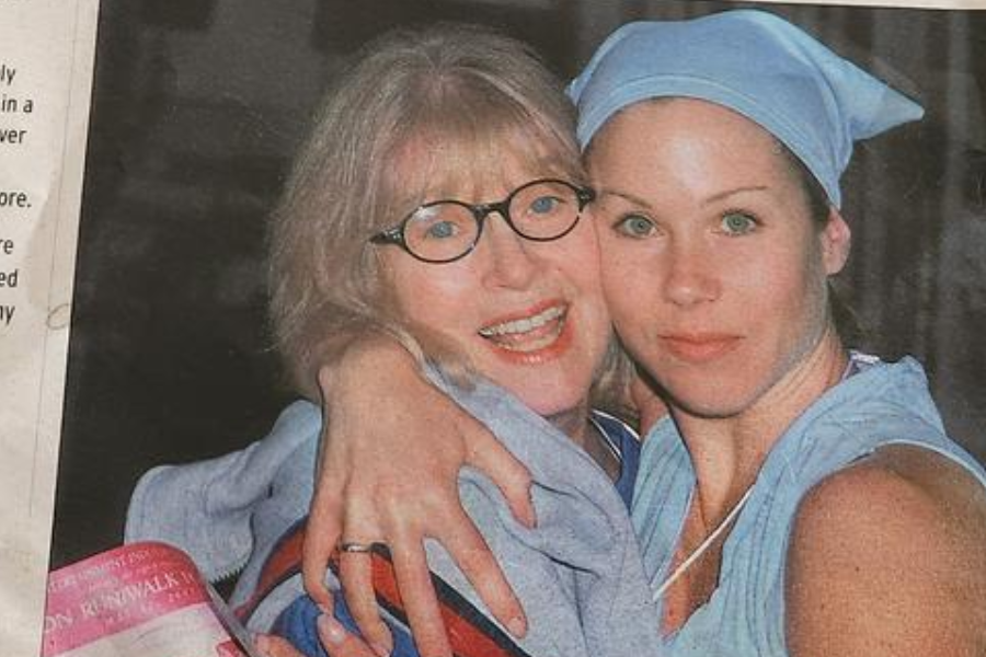 A younger woman in a light blue athletic outfit and headscarf hugs an older woman wearing glasses and a light jacket. They both smile warmly, appearing close and affectionate, with the older woman holding a pink event booklet.