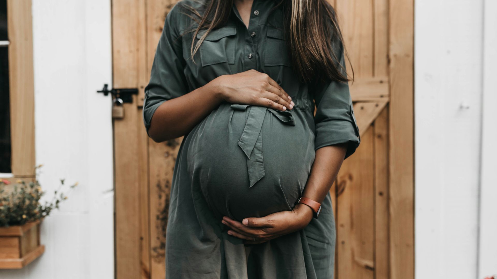 Pregnant woman standing near a door, gently holding her belly, expressing a moment of care or anticipation.