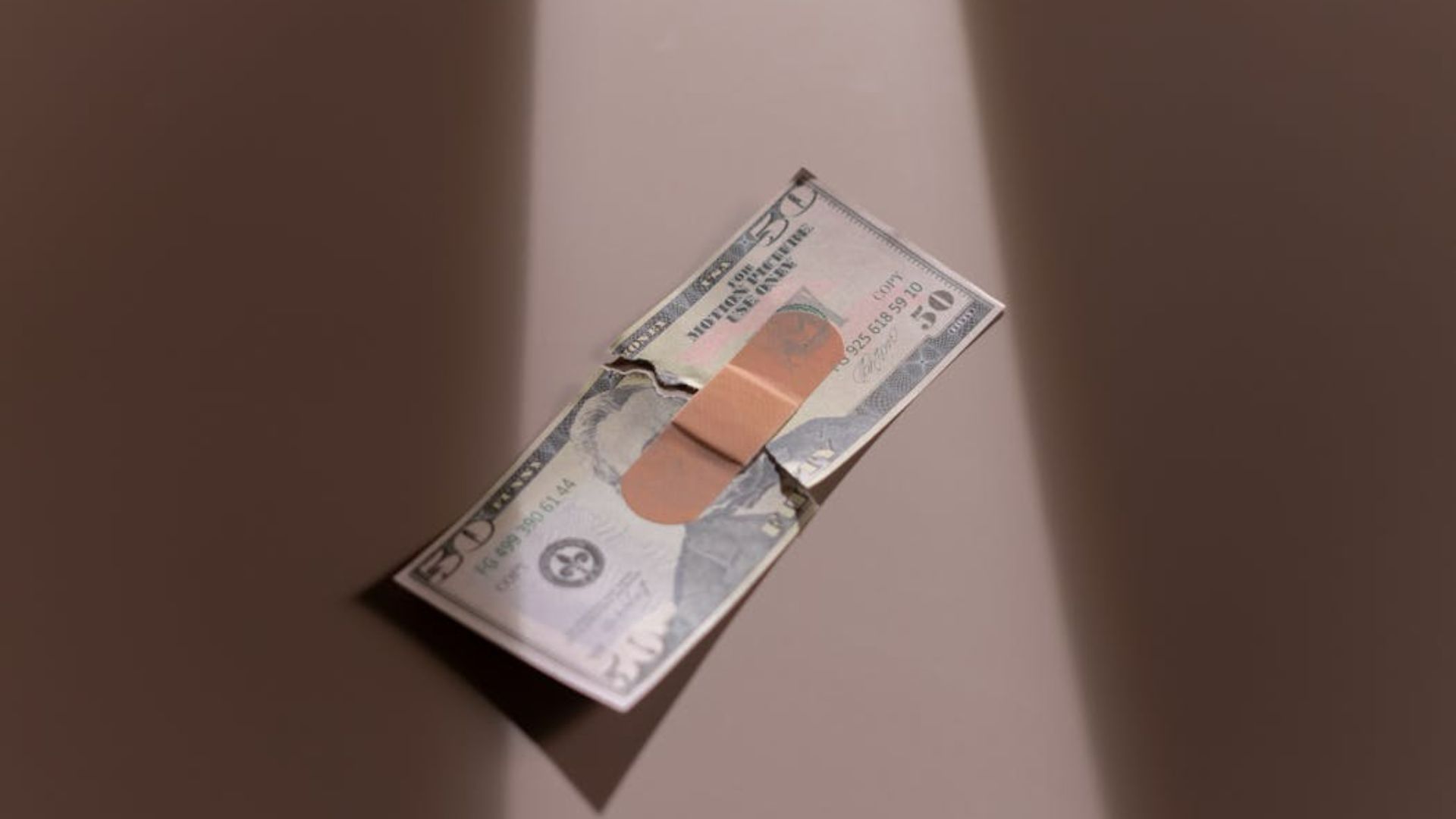 A torn 50-dollar bill is taped together with a bandage. It lies on a neutral surface, with shadows on each side, conveying a theme of repair and fragility.