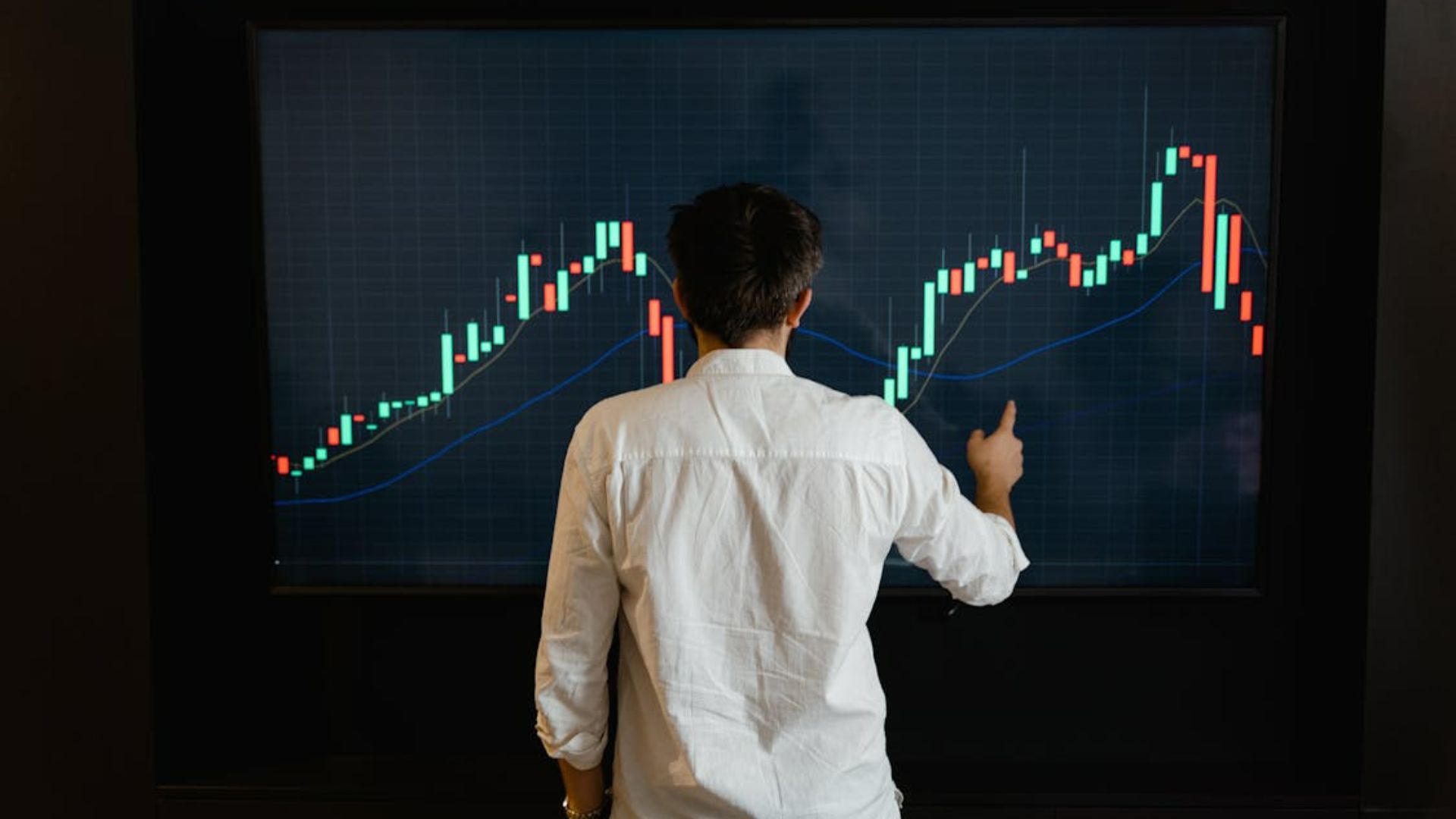 A person in a white shirt stands in front of a large screen displaying a fluctuating stock chart with green and red candlesticks, suggesting market analysis.