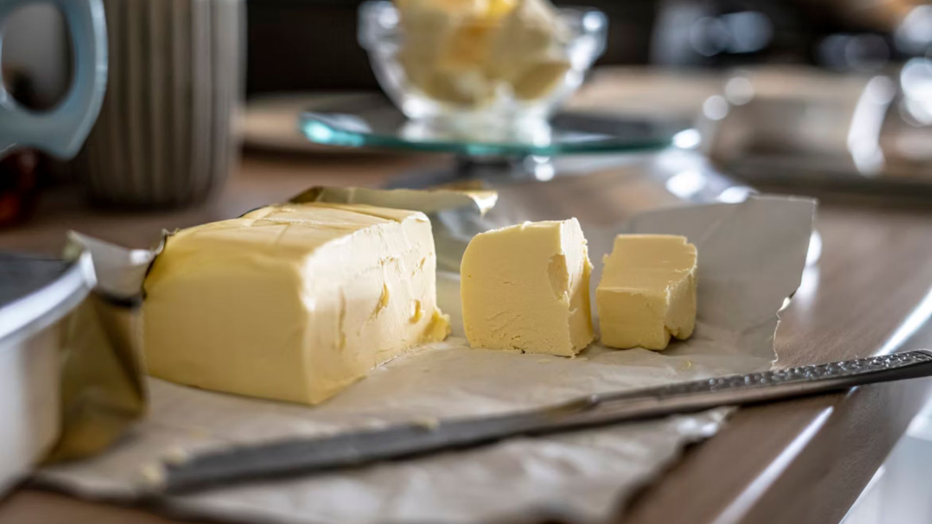 A block of butter with slices cut off rests on parchment paper on a kitchen counter. A knife lies beside it, with a soft focus background creating a warm, inviting ambiance.