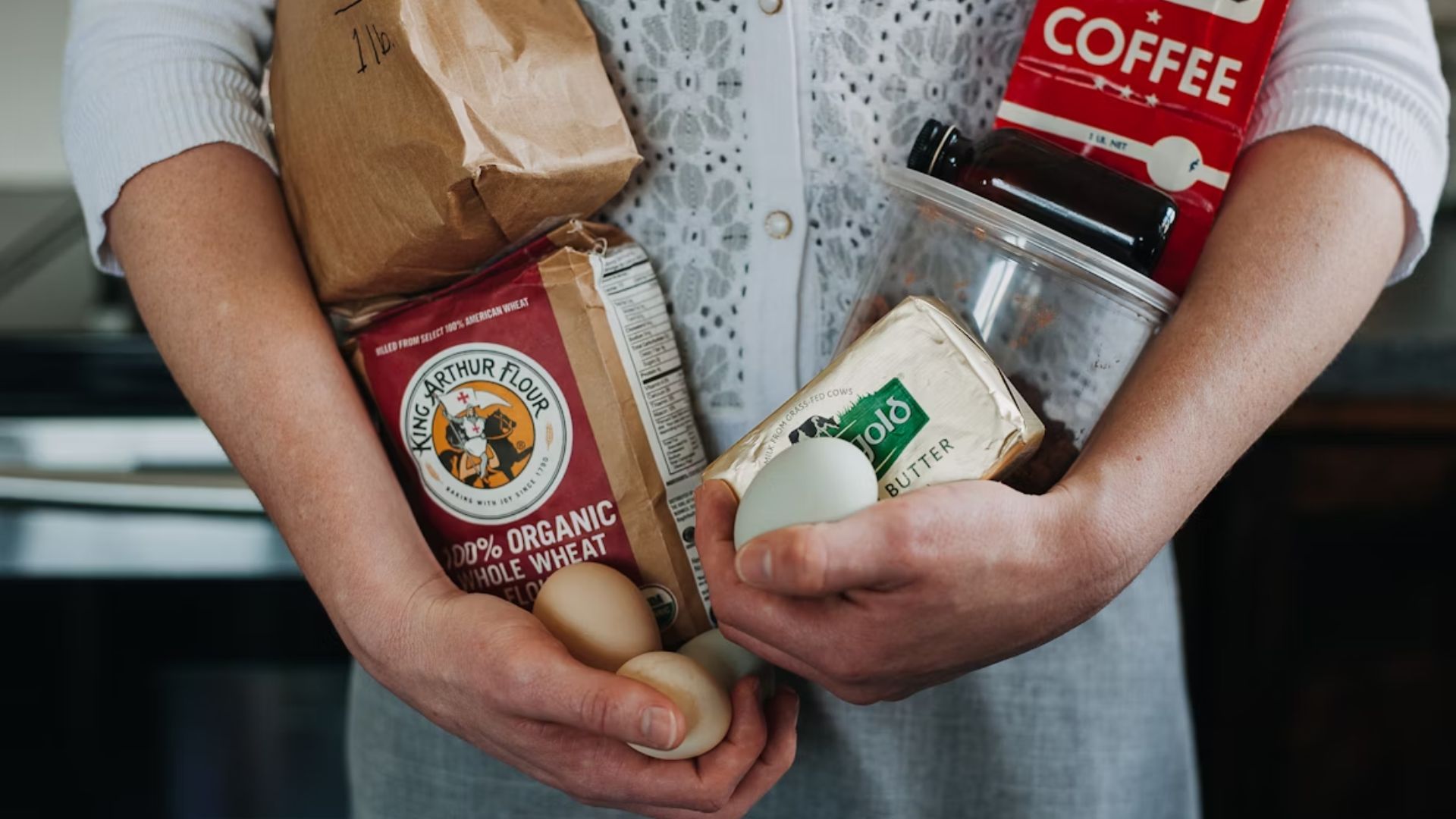 A person holds grocery items, including a bag of flour, coffee, butter, and eggs. The tone is homey, suggesting meal preparation or baking.