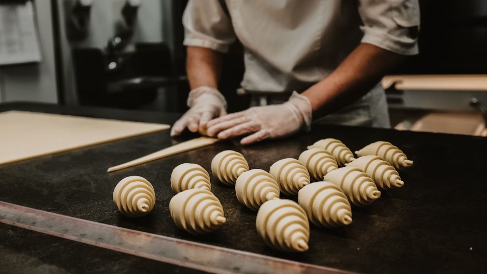 A baker in white uniform and gloves shapes dough on a black counter, preparing a neat row of spiral croissants. The setting exudes focus and artistry.