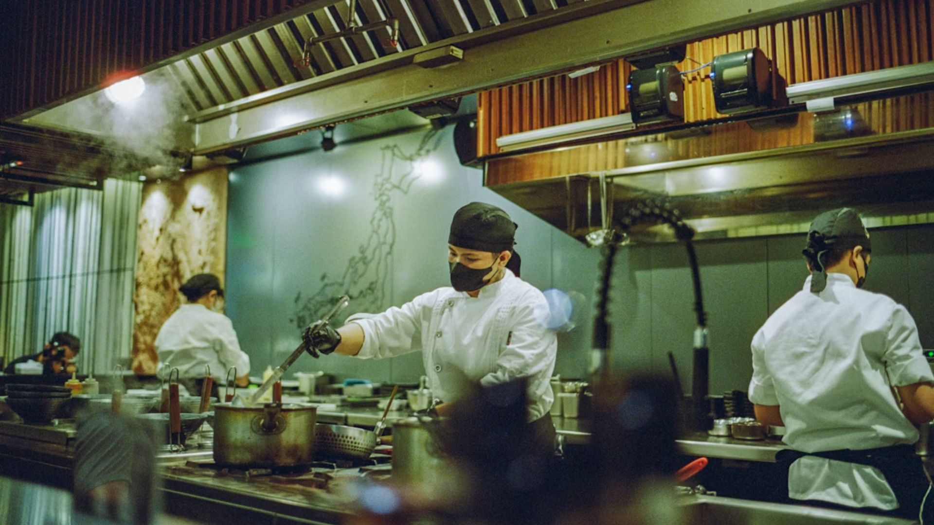 Three chefs in a bustling kitchen, wearing white uniforms and black masks. One chef is stirring a pot. Warm lighting creates a focused atmosphere.