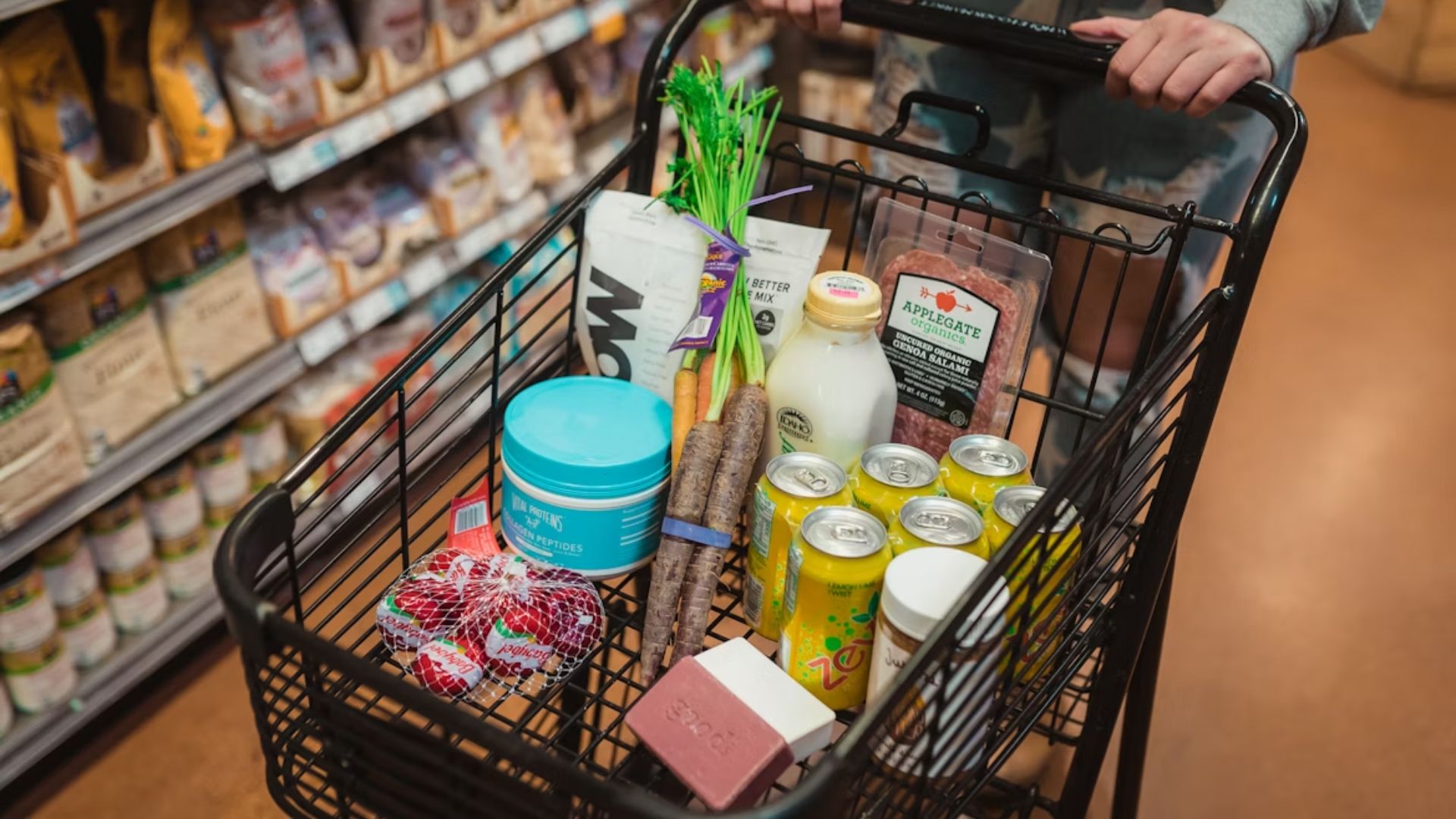A grocery cart in a store aisle contains fresh vegetables, milk, packaged meat, canned drinks, and toiletries, suggesting a balanced shopping haul.
