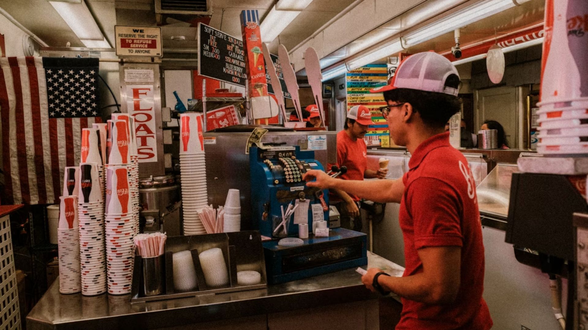 Vintage diner scene with two staff in red uniforms and caps, one using a blue register. Stacks of soda cups and an American flag create a nostalgic vibe.
