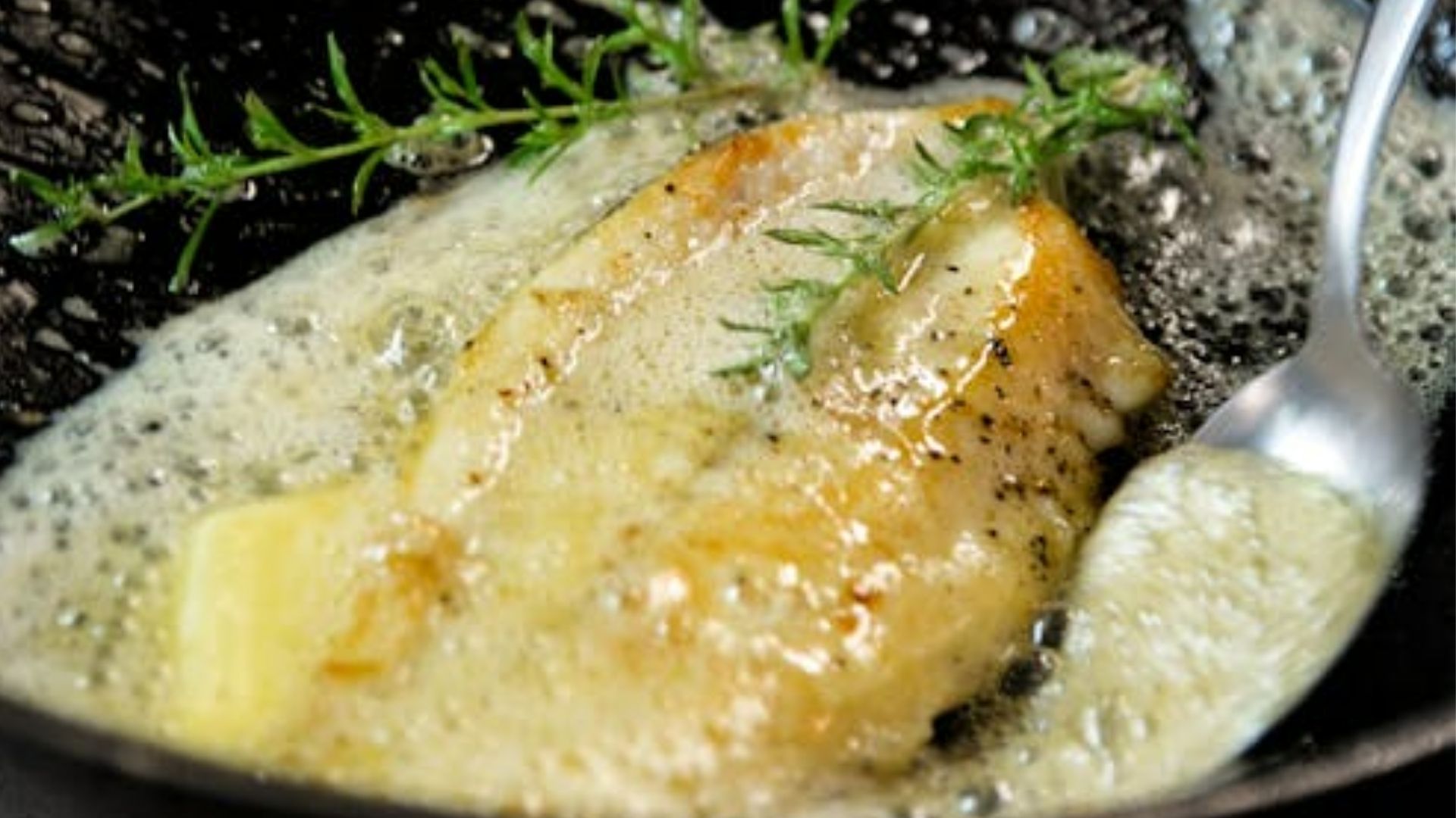 Golden-browned fish fillet sizzling in a pan with melted butter, fresh herbs, and a spoon, creating a rich and aromatic cooking scene.